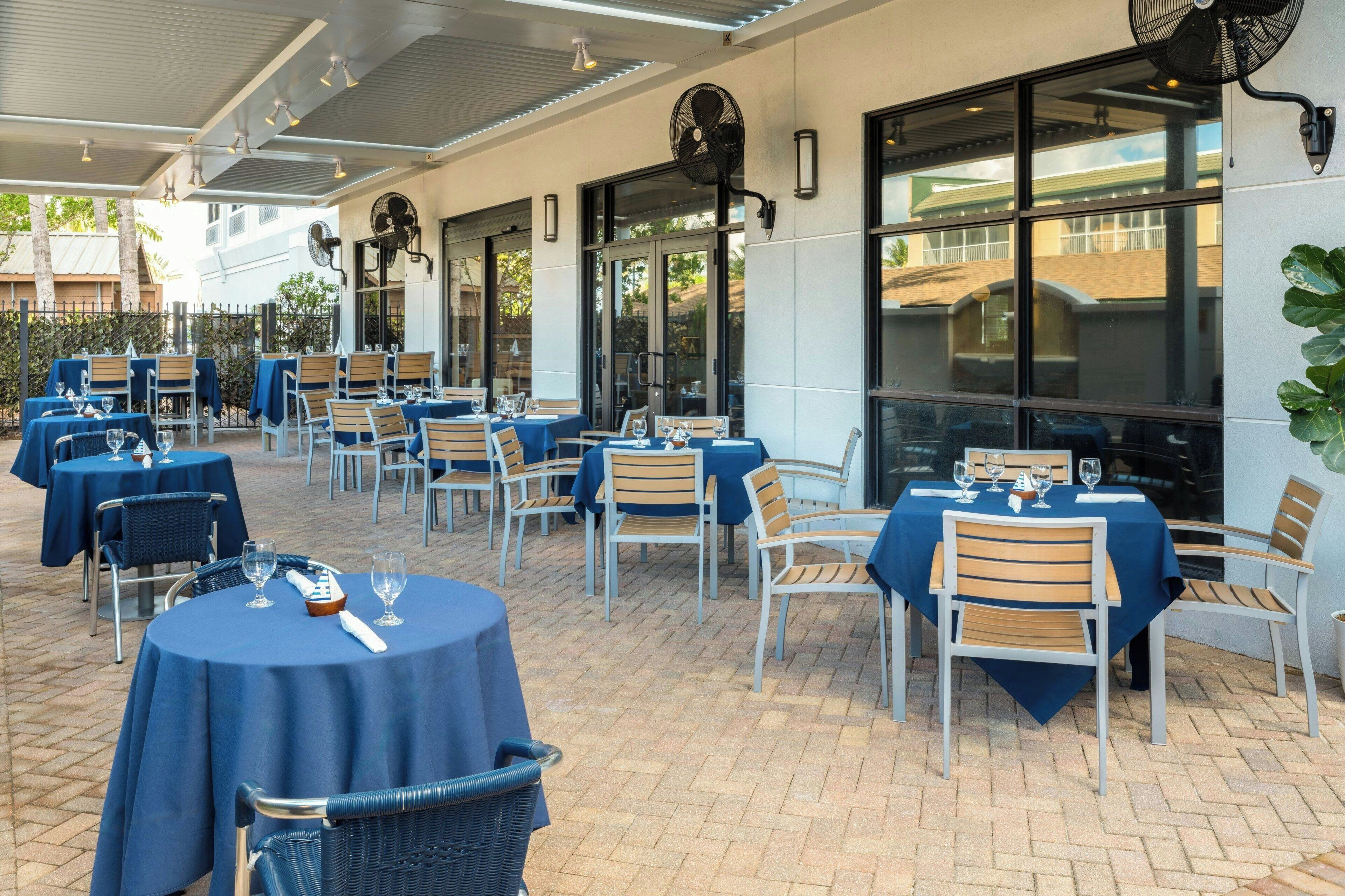 Eine Terrasse im Freien mit runden Tischen und Stühlen, bedeckt mit blauen Tischdecken, und mit Deckenventilatoren ausgestattet.
