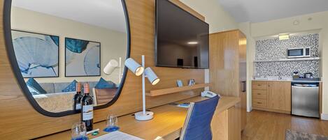 Una habitación de hotel moderna con un escritorio de madera, un espejo grande, un televisor de pantalla plana, una zona de cocina con microondas y una mesa de comedor.