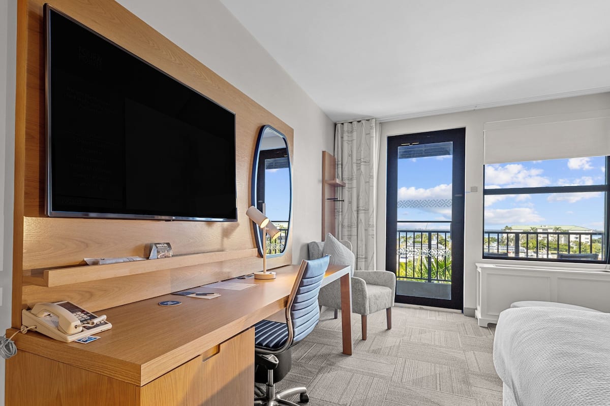 A hotel room with a wooden desk, a flat-screen TV, a chair, a bed, and a balcony with a view.