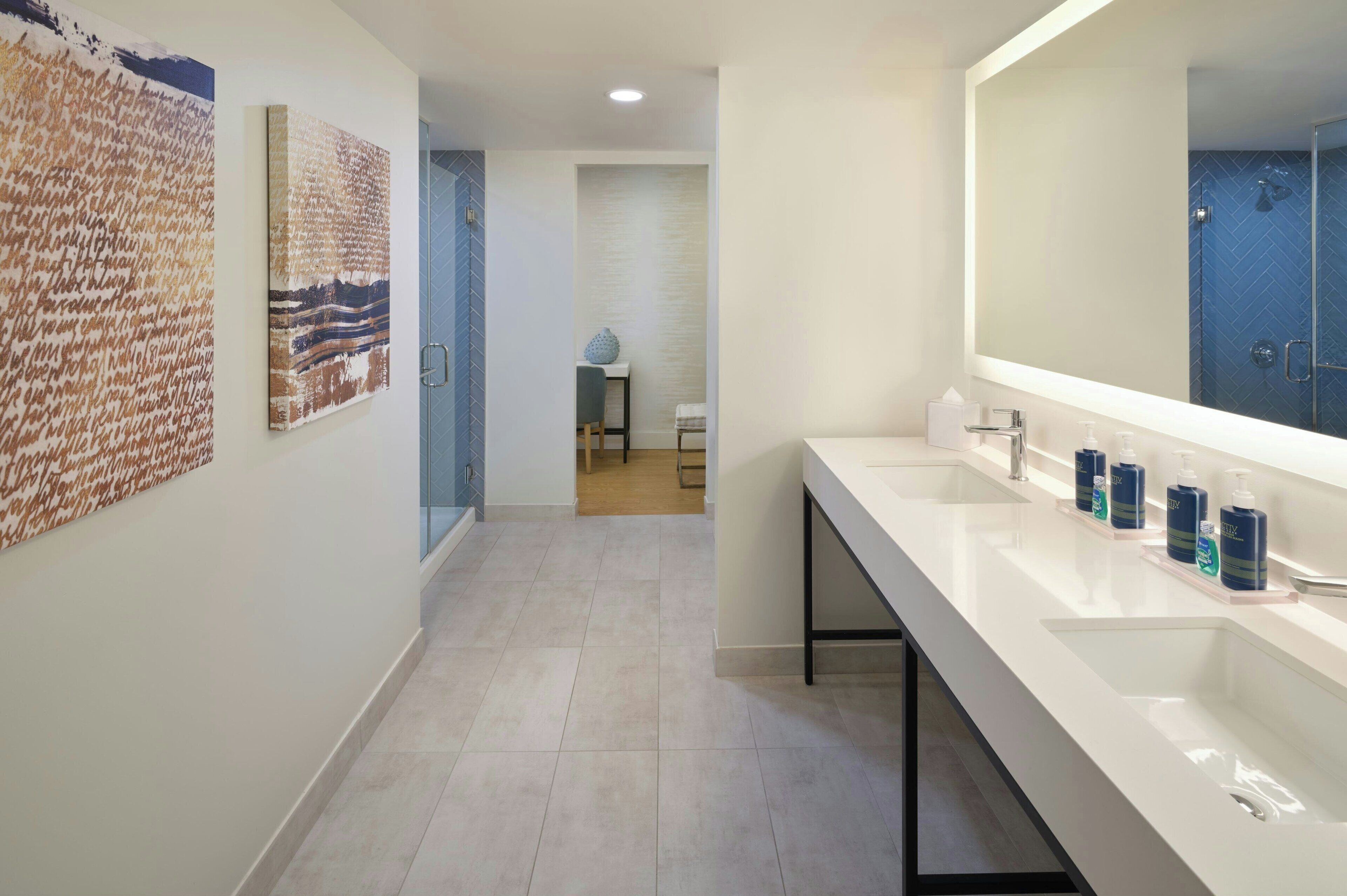 A modern bathroom with a long white sink, a large mirror, and two abstract paintings on the wall.