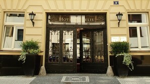 A hotel facade with a symmetrical design, featuring a central entrance with ornate glass doors, a sign above the entrance, and two potted plants on either side. - Hotel Angelis (Prague)