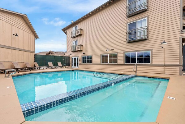 A rectangular swimming pool with a tiled edge, surrounded by lounge chairs and a building with balconies.
