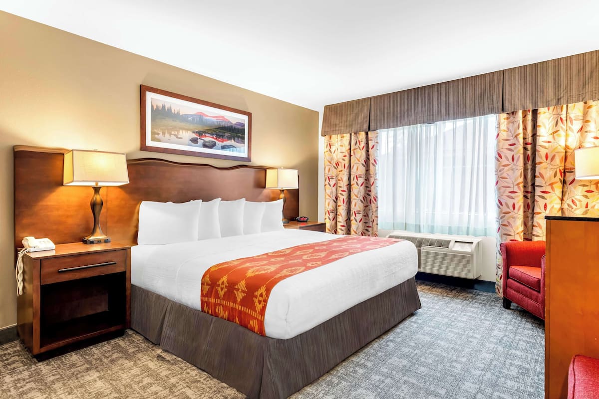 Een hotelkamer met een groot bed, twee nachtkastjes met lampen, een rode fauteuil, een bureau en een raam met gordijnen.