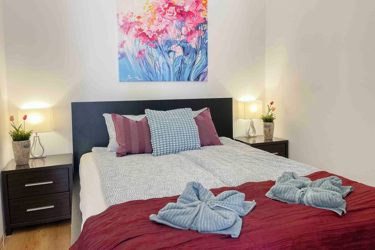 A neatly made bed with a red blanket and two towels, a dark headboard, and a wall-mounted painting of flowers.