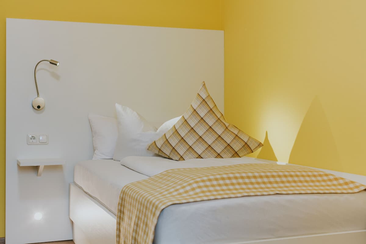 A neatly made bed with a checkered blanket, a bedside lamp, and a pillow with a plaid pattern.