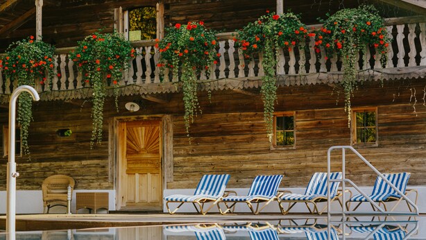 A wooden cabin with a pool, blue-striped lounge chairs, and hanging flower baskets. - Hotel Drei Quellen Therme (Bad Griesbach im Rottal)