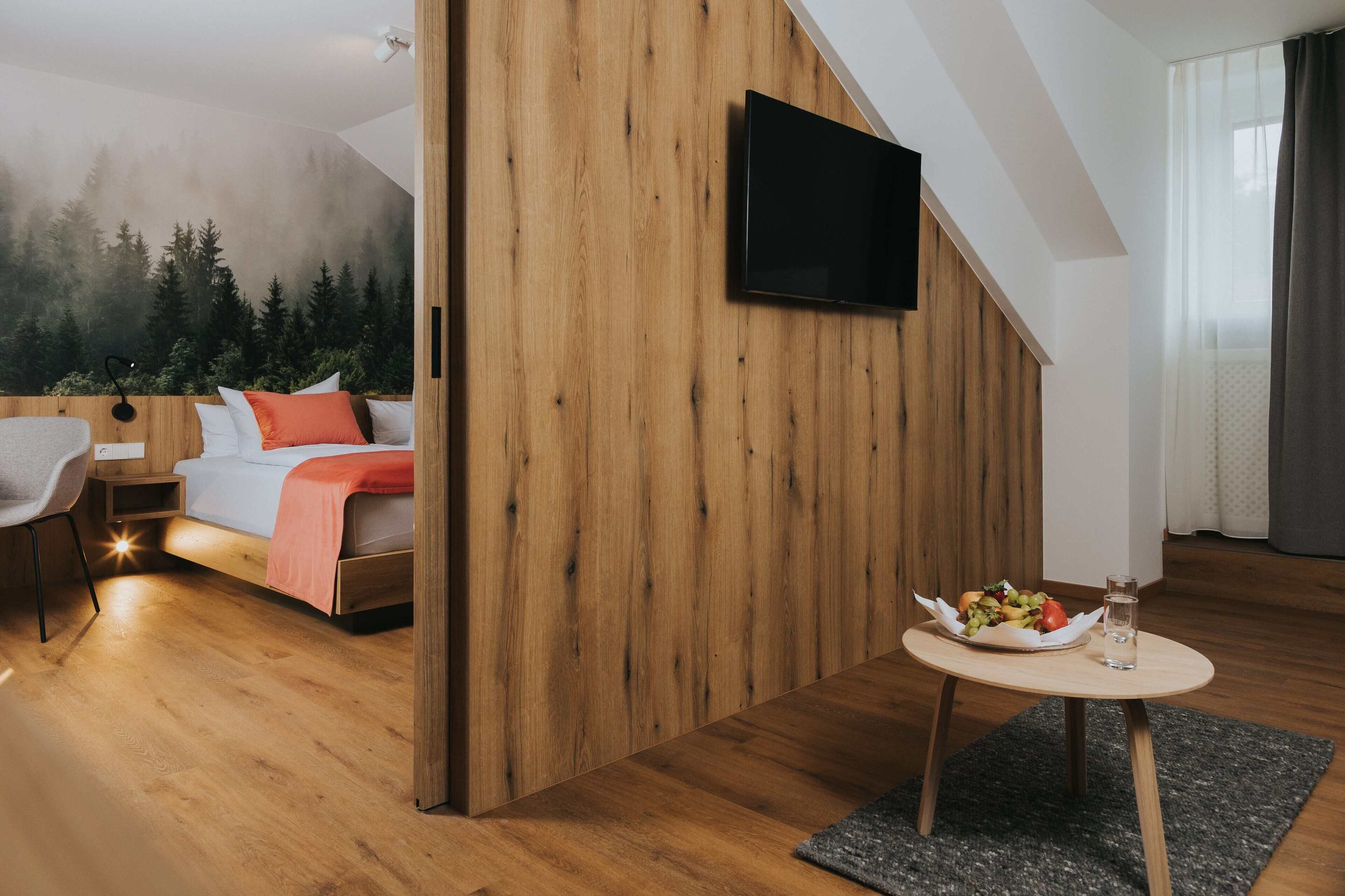 A modern room with a bed, a TV mounted on a wooden wall, a small table with a fruit bowl and glass, and a cozy chair.