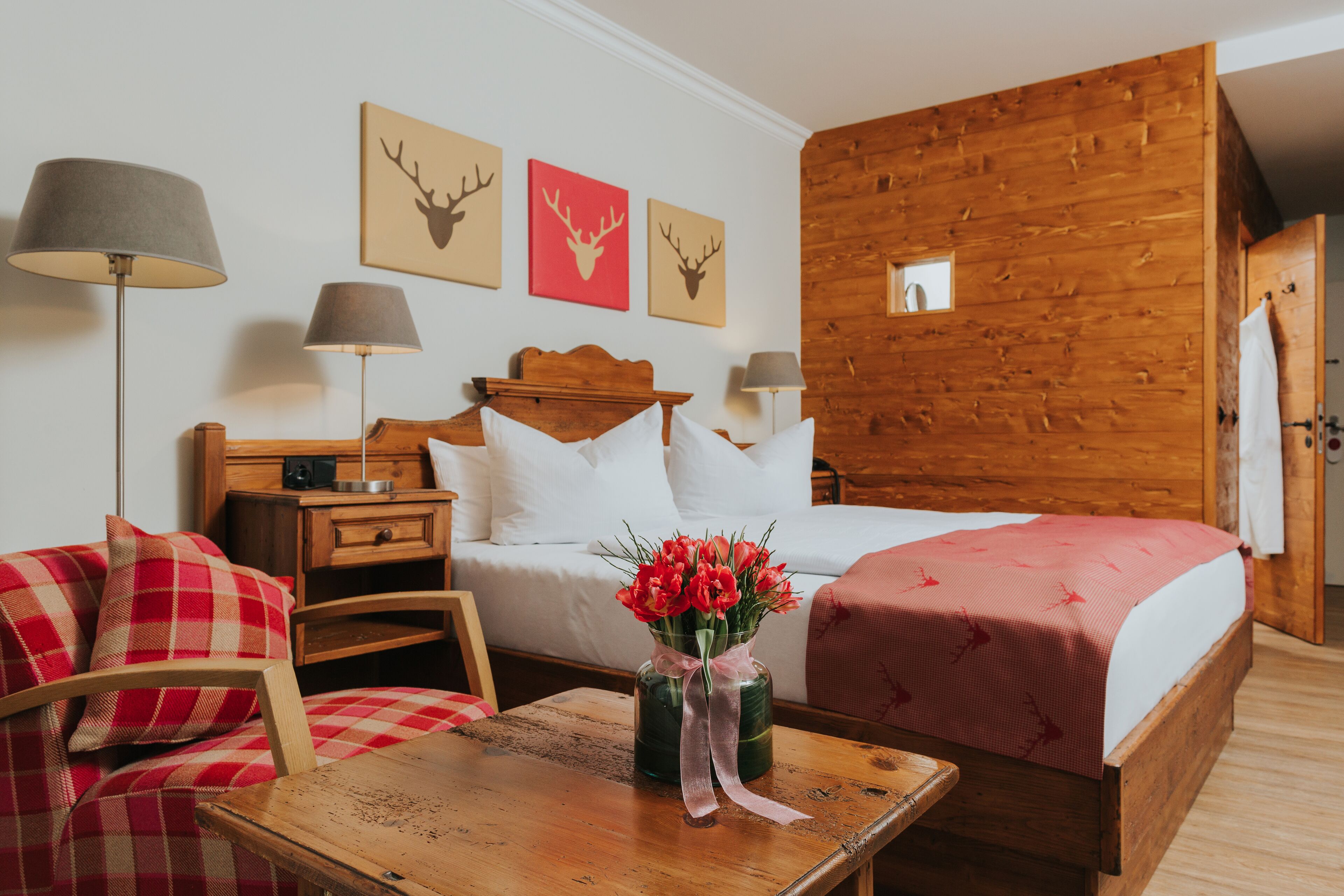 A wooden cabin-style room with a bed, a wooden nightstand, a chair, a table with a vase of flowers, and wall-mounted lamps.