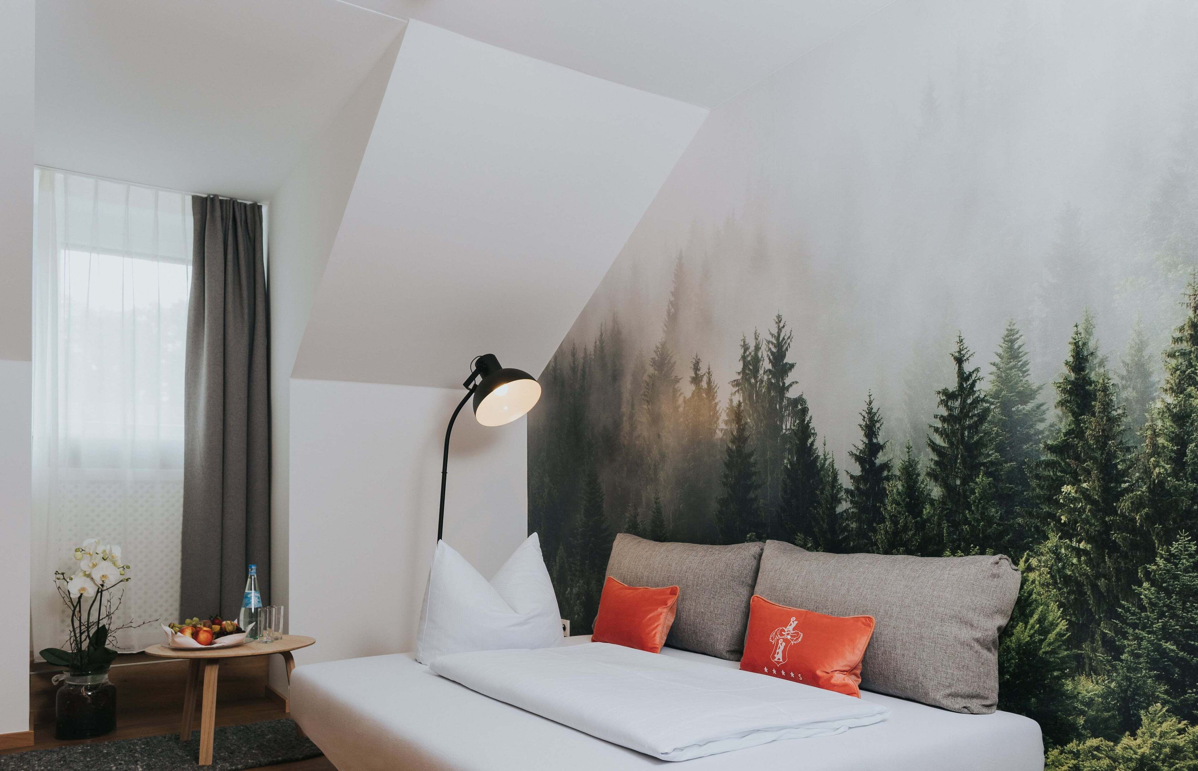 A modern hotel room with a bed, a nightstand, a lamp, and a mural of a forest.