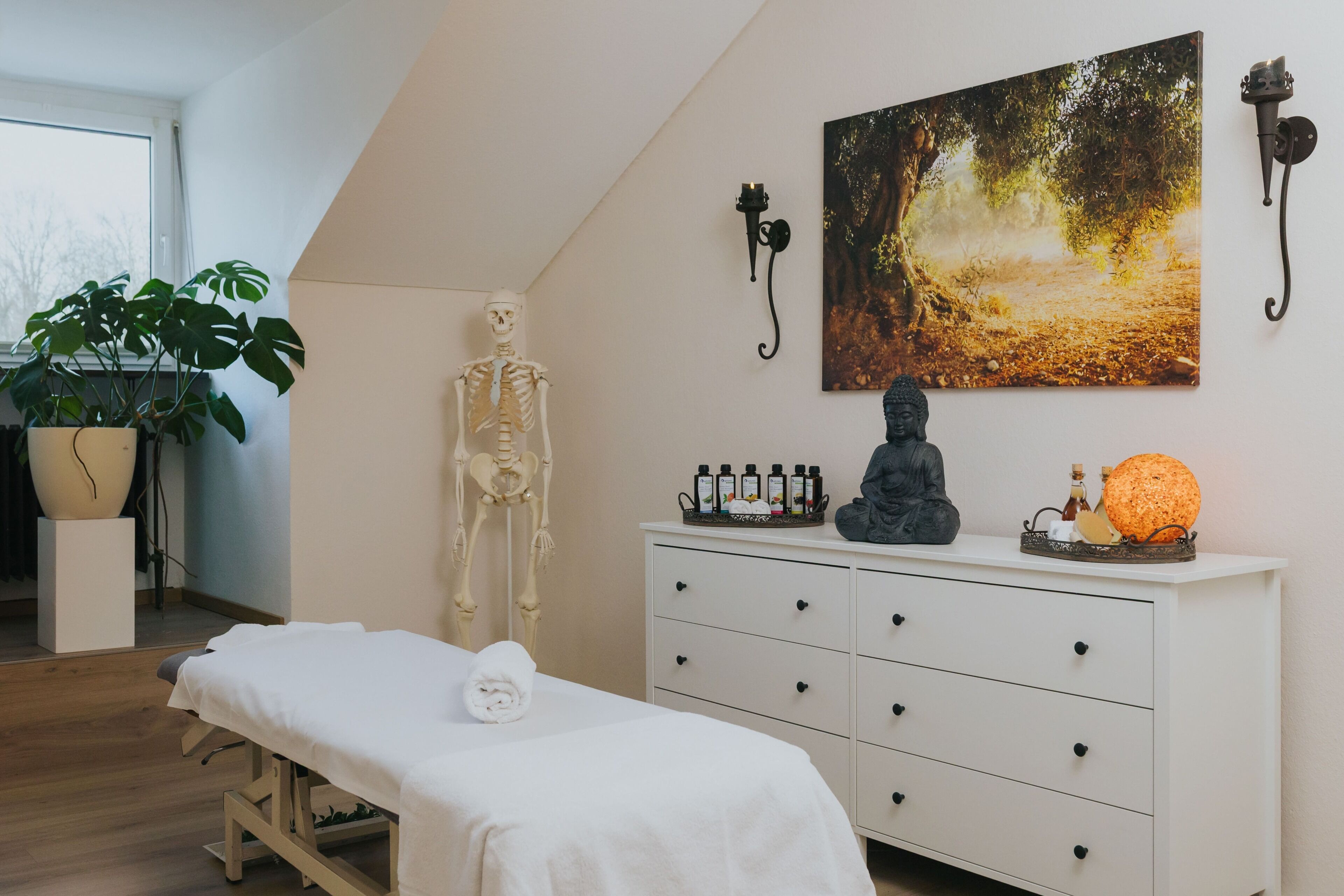 A room with a massage table, a skeleton, a Buddha statue, a painting, and a potted plant.