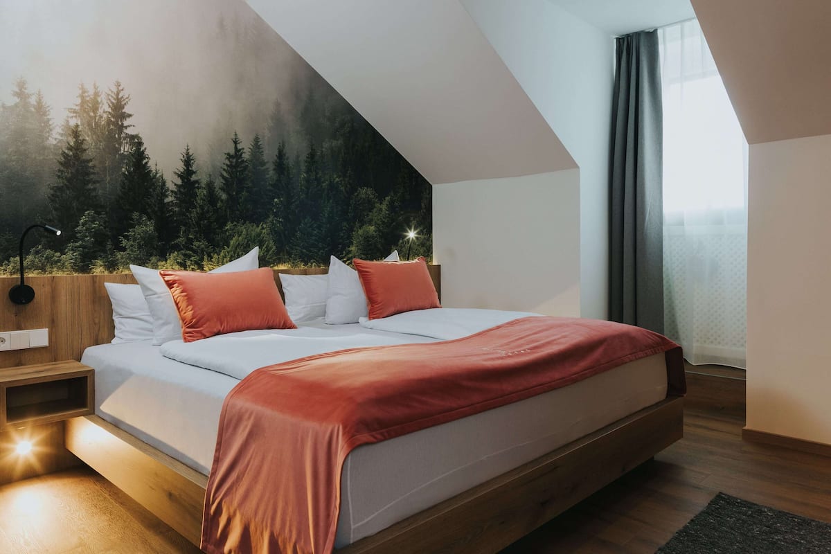 A modern bedroom with a sloped ceiling, a bed with white and red bedding, a nightstand, and a wall mural of a forest.