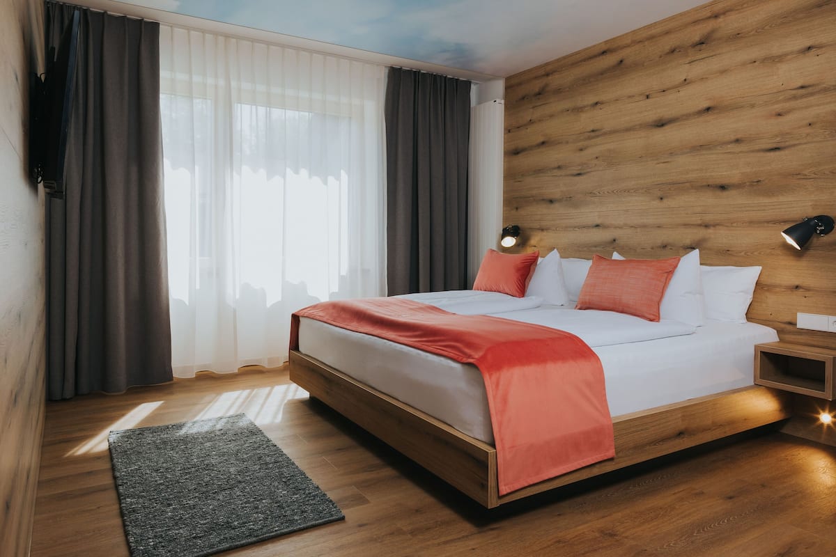 A modern bedroom with a wooden headboard, a large bed with white and orange bedding, a bedside table with a lamp, and a view of the city through sheer curtains.