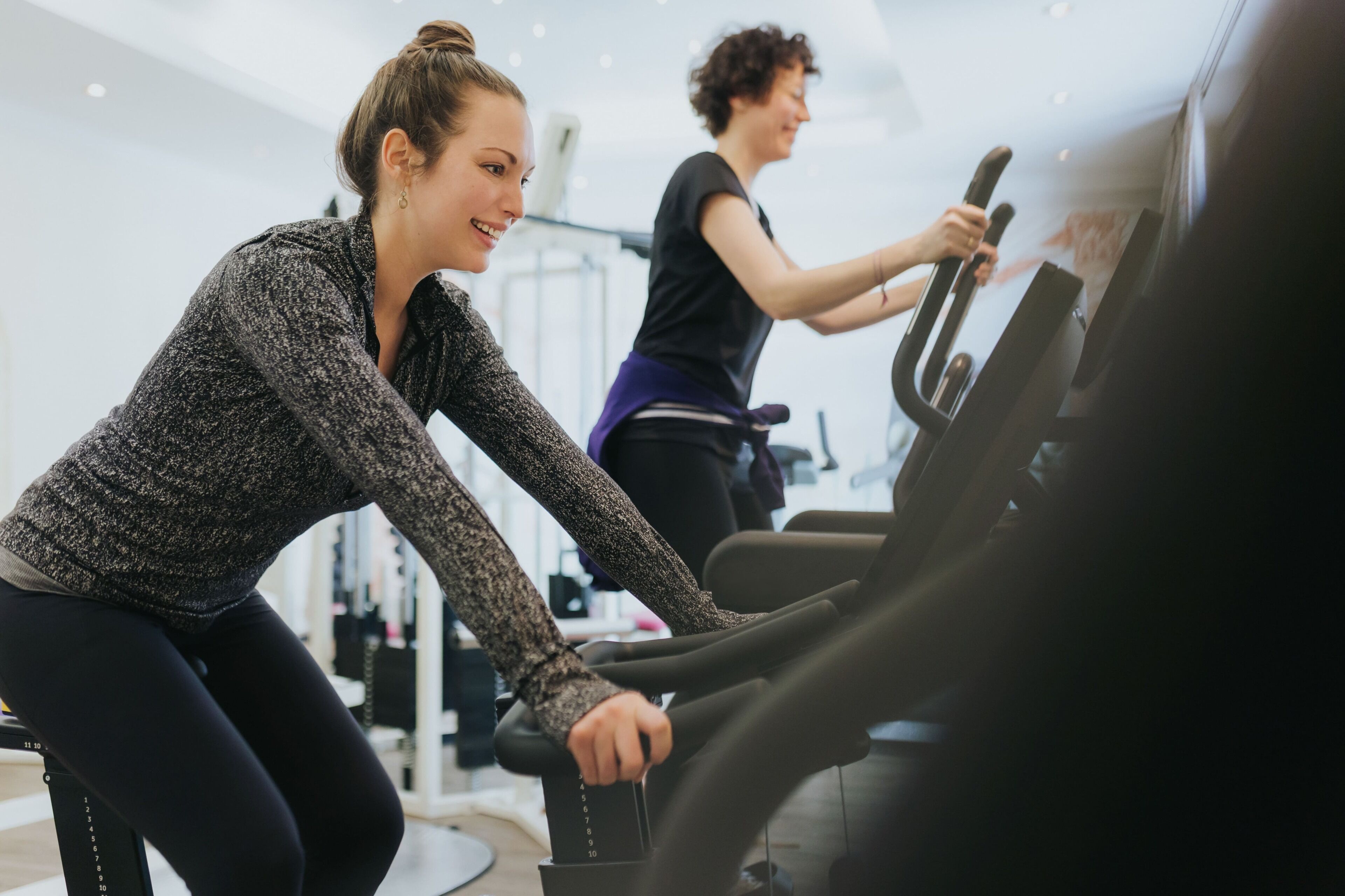 Two individuals using stationary bikes in a gym with exercise equipment and mirrors.
