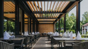 An outdoor dining area with wooden tables and chairs, covered by a wooden pergola, and large windows offering views of the surrounding greenery. - Hotel Drei Quellen Therme (Bad Griesbach im Rottal)