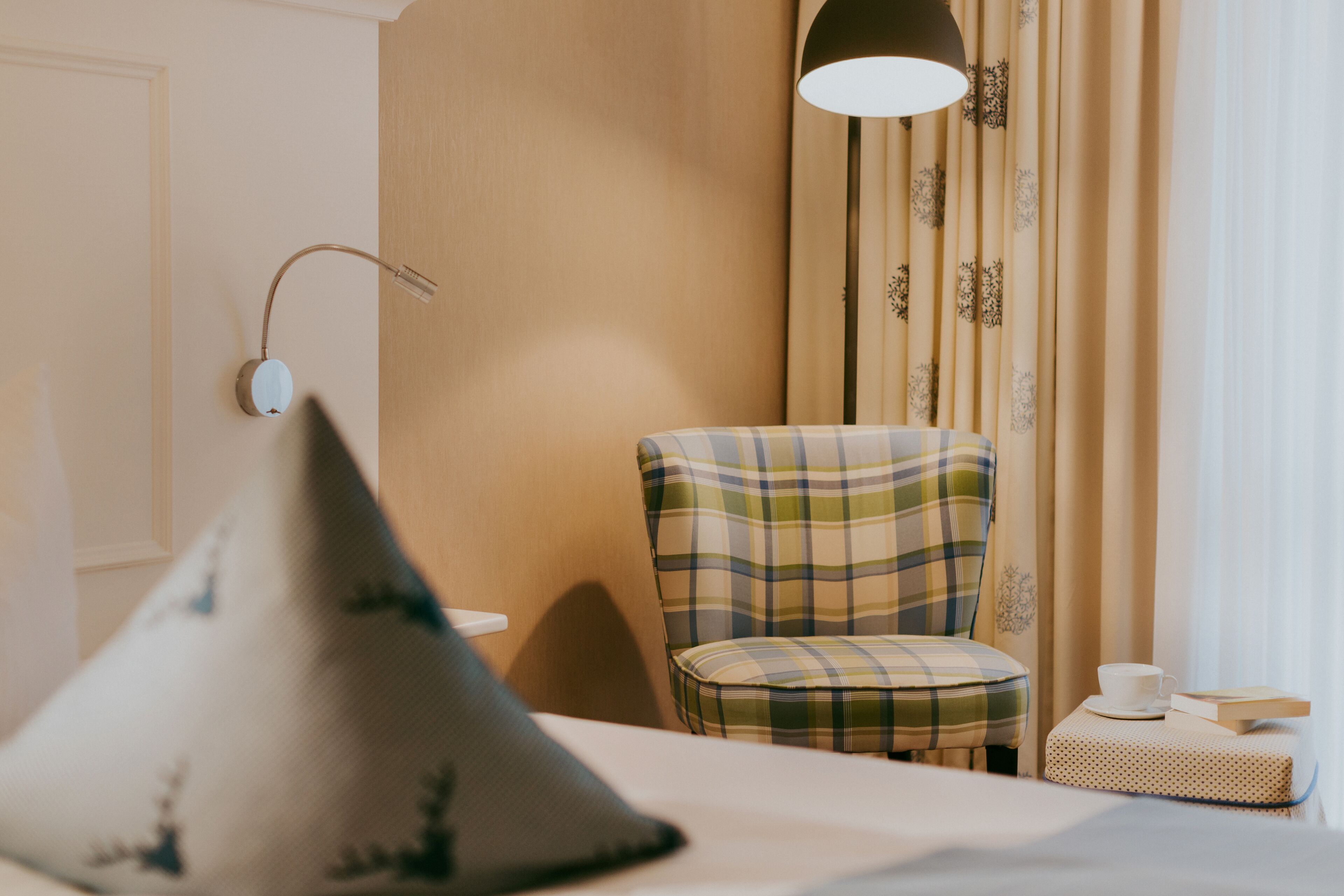 Une chambre d’hôtel avec un lit, un fauteuil à motifs écossais, une petite table avec une tasse et un livre, et une lampe fixée au mur.