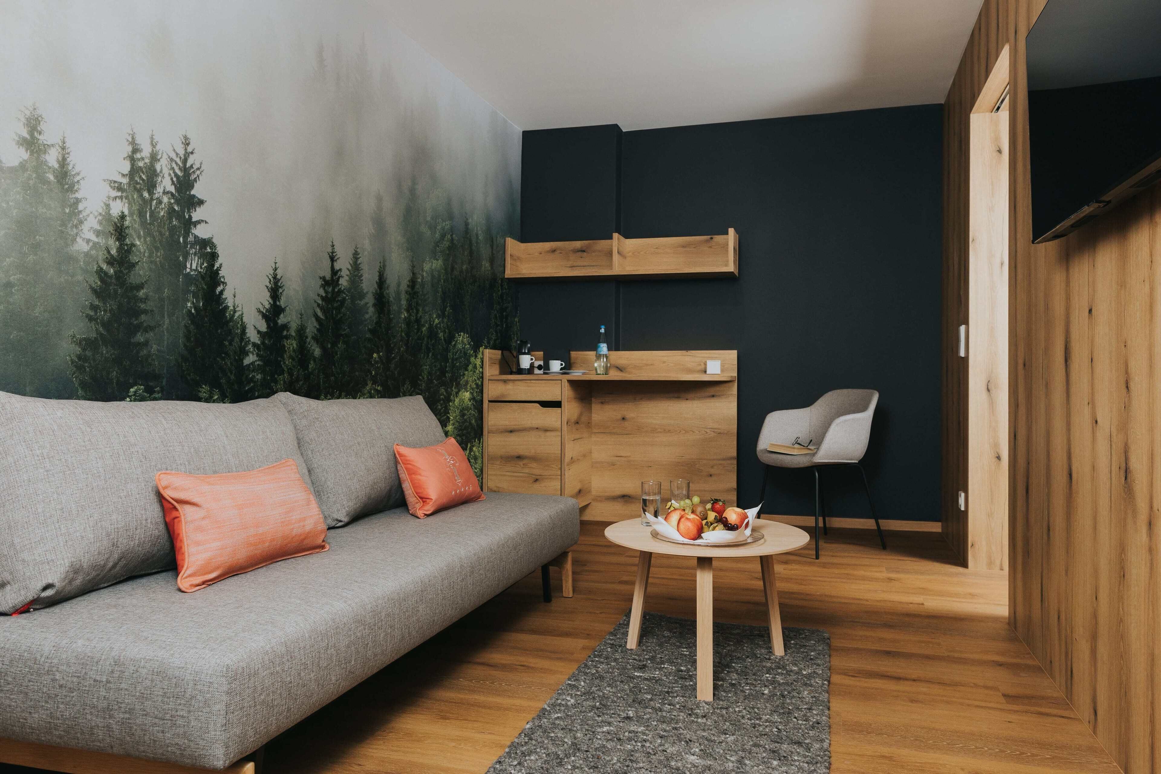 A modern living room with a grey sofa, orange cushions, a small round table with a fruit basket, and a wall-mounted wooden shelf.