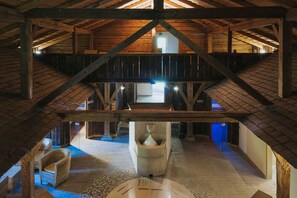 Un interior rústico con techo alto, vigas de madera, una escalera central y una zona de estar con una mesa redonda.
