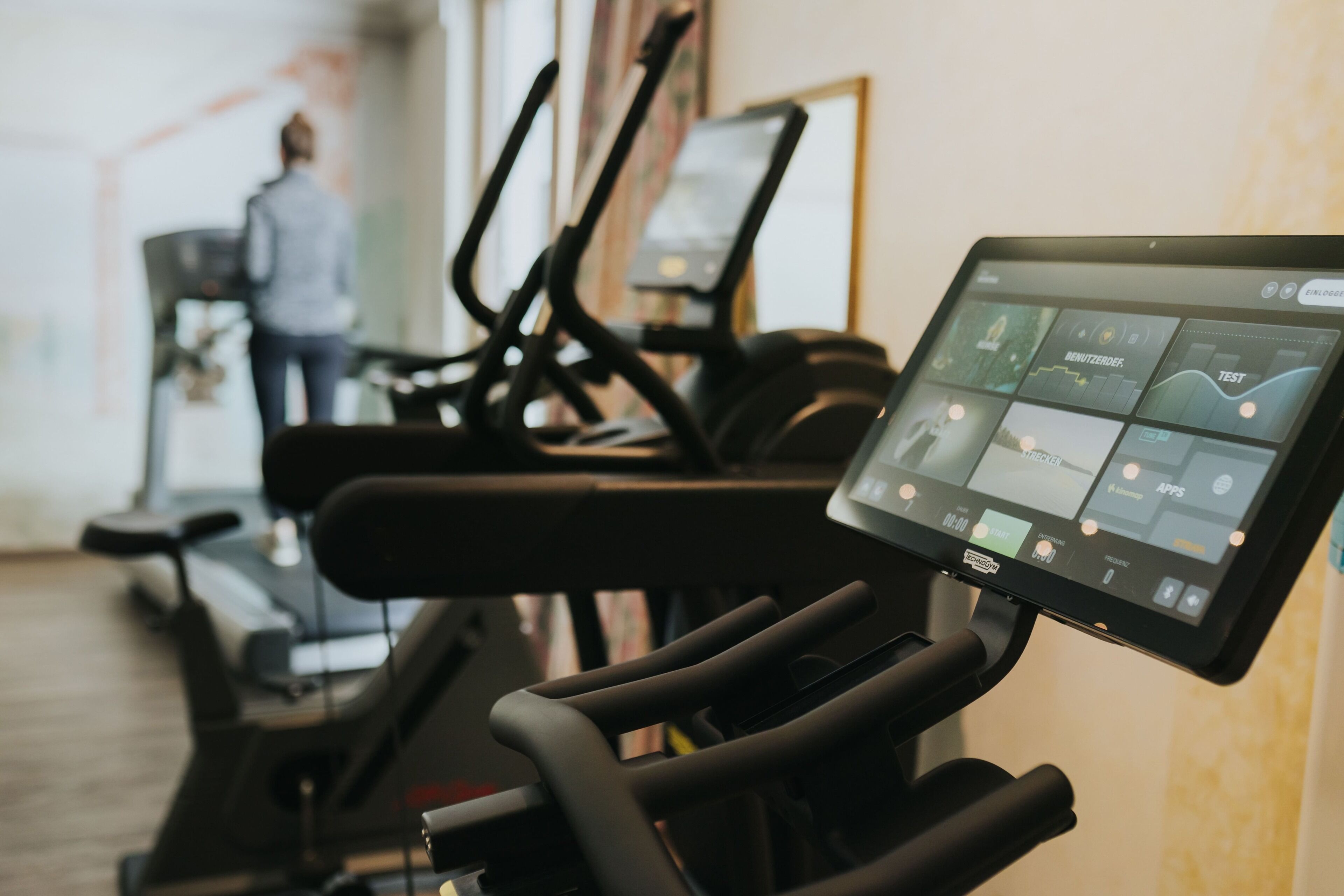 A treadmill with a digital display, a wall-mounted screen, and exercise equipment in a gym setting.