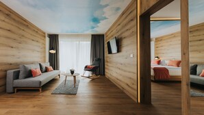 A modern living room with wooden panel walls, a grey sofa, a small round table, and a TV mounted on the wall. - Hotel Drei Quellen Therme (Bad Griesbach im Rottal)