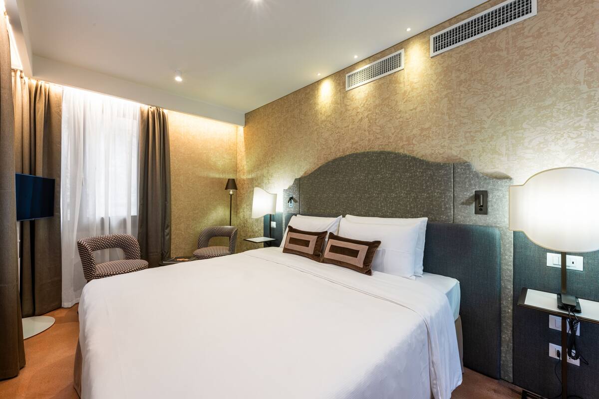 A hotel room with a large bed, a chair, a TV, a lamp, and a wall-mounted heater.