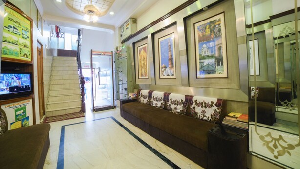Una hall d'albergo con una reception, una scala, un divano con cuscini decorativi, quadri appesi al muro e un televisore che mostra una veduta cittadina.