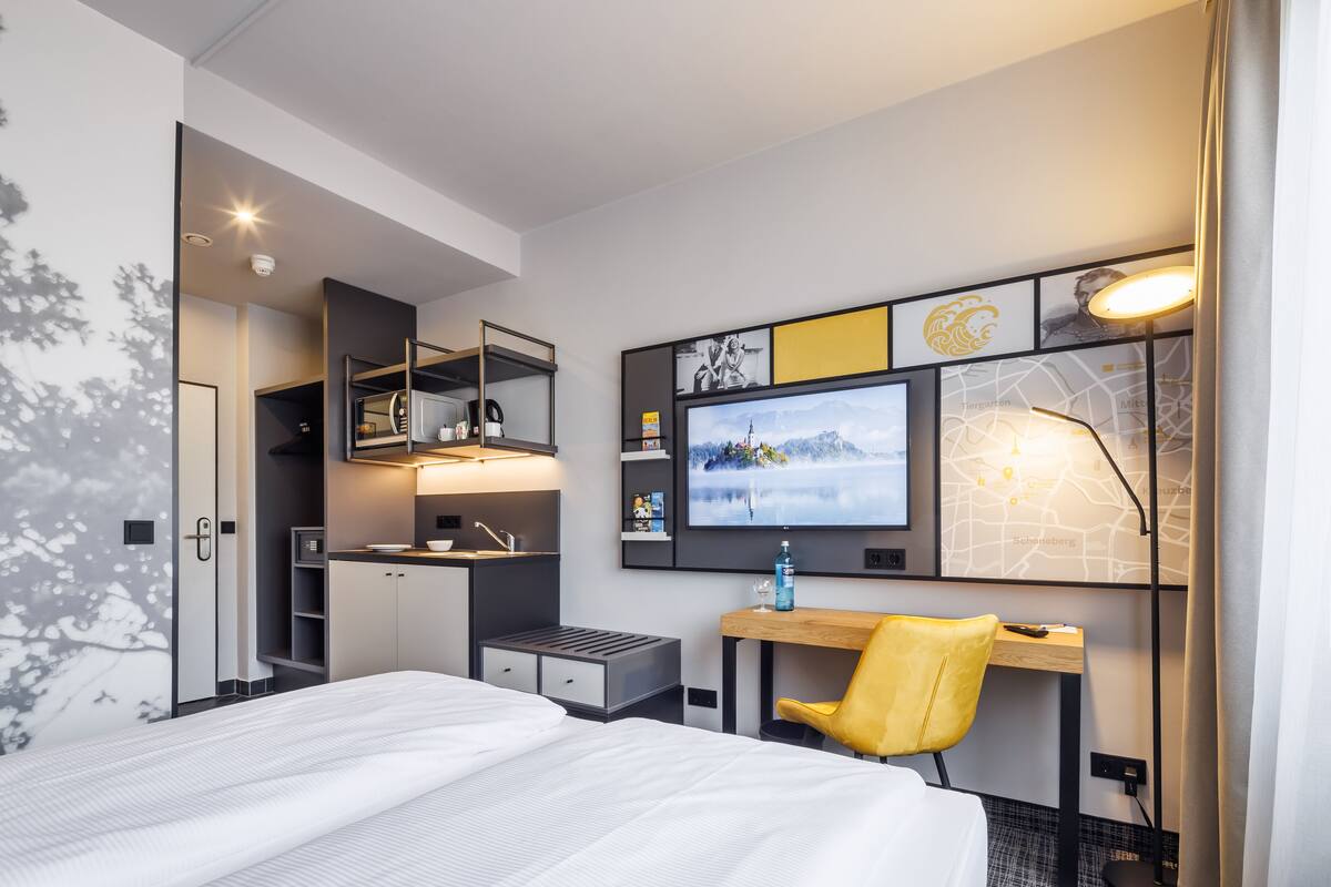 A modern hotel room with a bed, a desk with a chair, a TV, and a map on the wall.