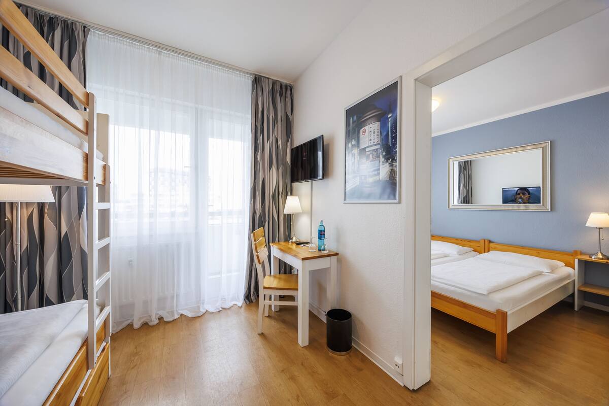 A hotel room with a bunk bed, a desk with a chair, a television, a bed, a mirror, and a window with curtains.