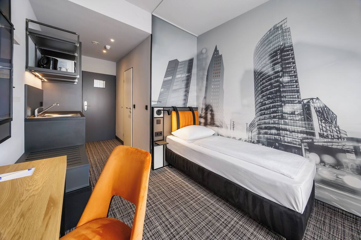 A hotel room with a bed, a desk, a chair, a small kitchenette, and a cityscape mural on the wall.