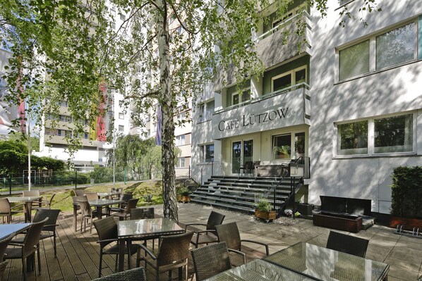 An outdoor cafe area with tables and chairs, a staircase leading to a building with a sign reading "CAFE LÜTZOW".