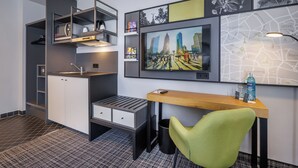 Kompakti hotellihuone, jossa on pieni keittiönurkkaus, työpöytä tuolilla, televisio ja seinällä oleva kartta.