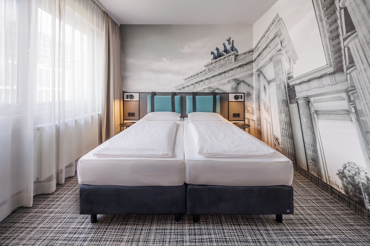 A hotel room with a large bed, two bedside tables, and a mural of a classical building.