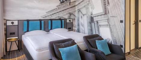 A hotel room with two beds, a large mural of a building, and two armchairs with blue cushions.