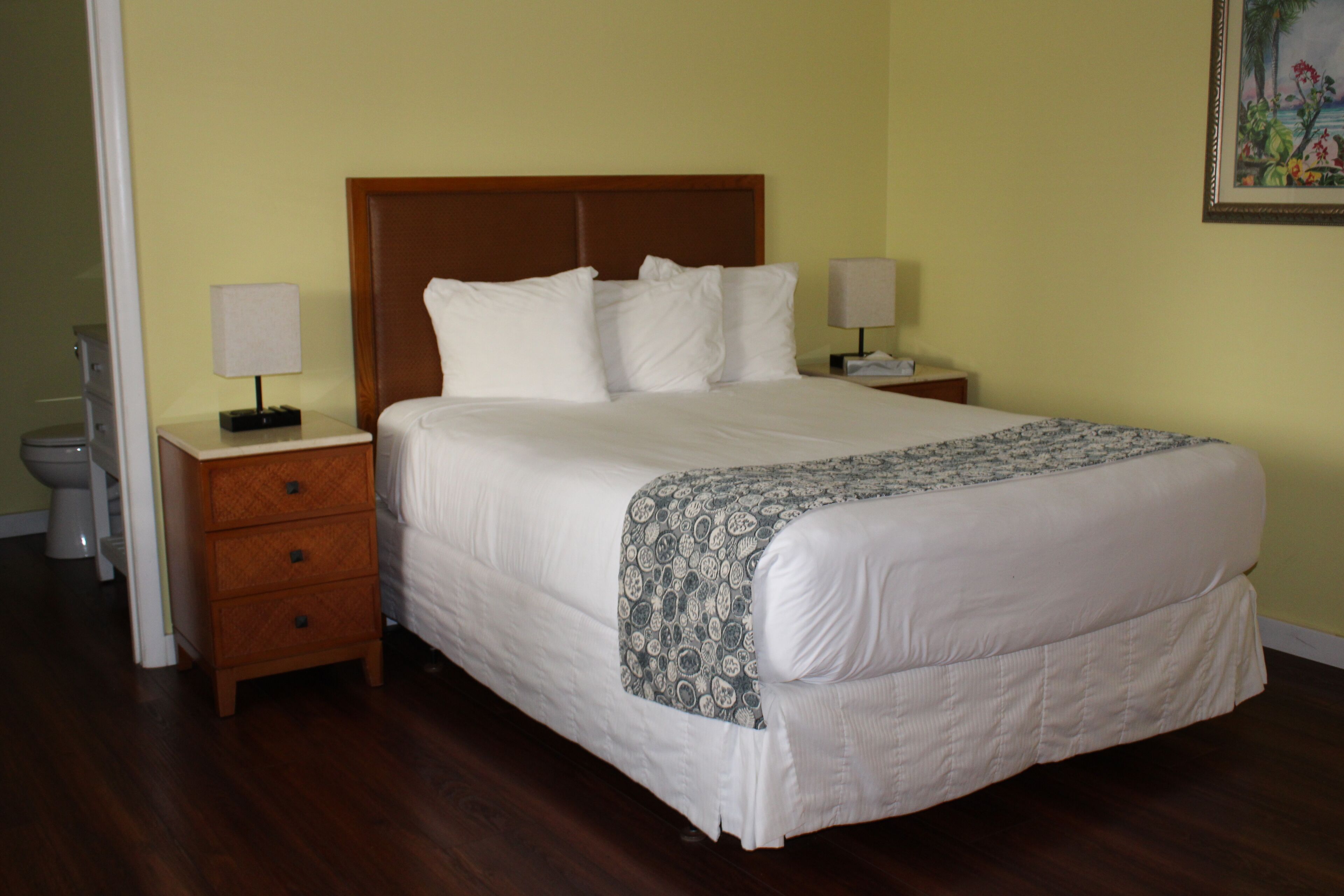 A neatly made bed with a patterned throw, a wooden nightstand with a lamp, and a small drawer.