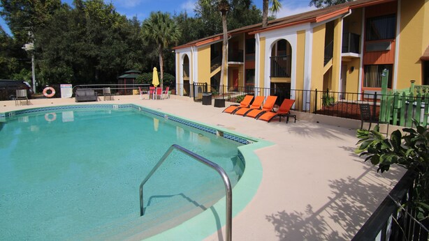 Zona de piscina con una piscina azul cristalina, sillas de descanso color naranja y un edificio con fachada amarilla y balcones.