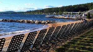 Uma praia com fileiras de cadeiras de metal na orla, um molhe rochoso e um barco atracado à direita.