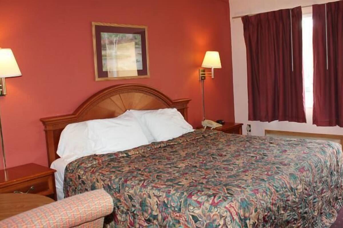 Quarto de hotel com cama de madeira, duas luminárias de cabeceira, uma janela grande com cortinas e um quadro pendurado na parede.