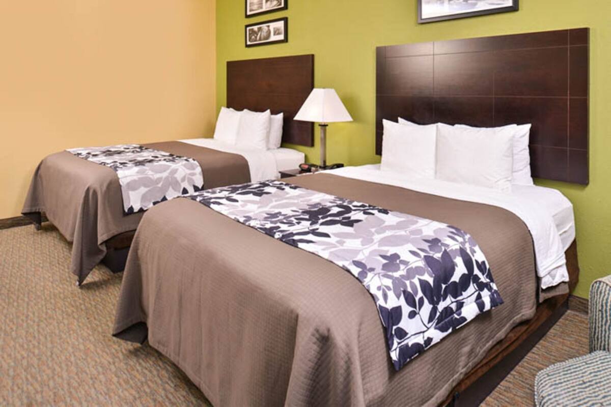 A hotel room with two beds, a headboard, a lamp, and a chair.