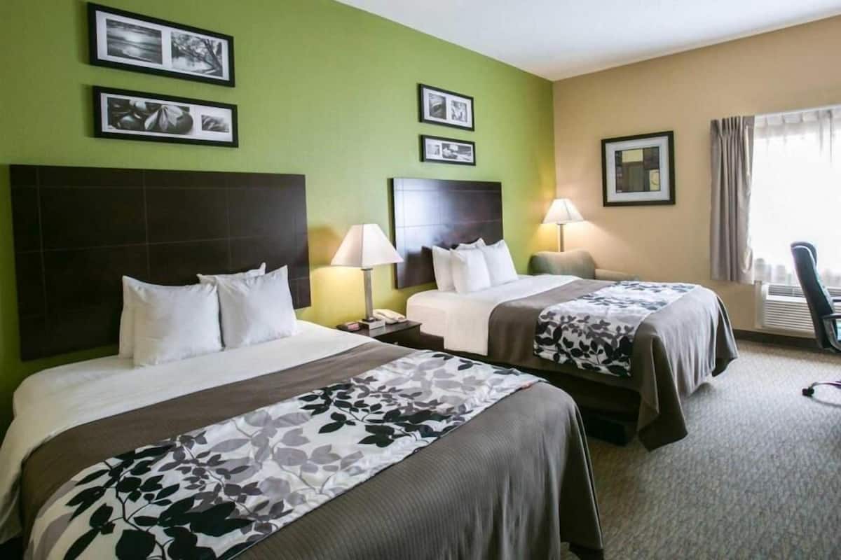 A hotel room with two beds, a nightstand, a lamp, a chair, and a window with curtains.