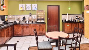 A compact kitchen with wooden cabinets, a round dining table with chairs, and a green wall with artwork.