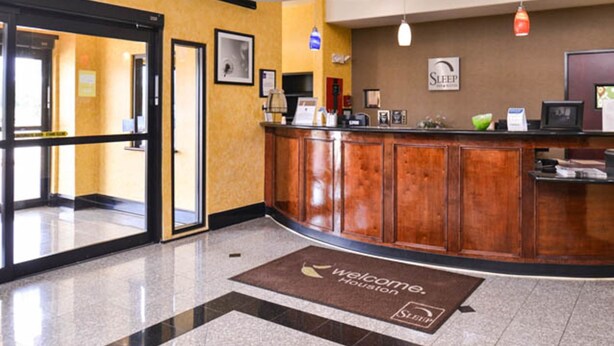 A hotel reception area with a wooden reception desk, a 'Welcome Houston' mat, and an 'EXIT' sign.