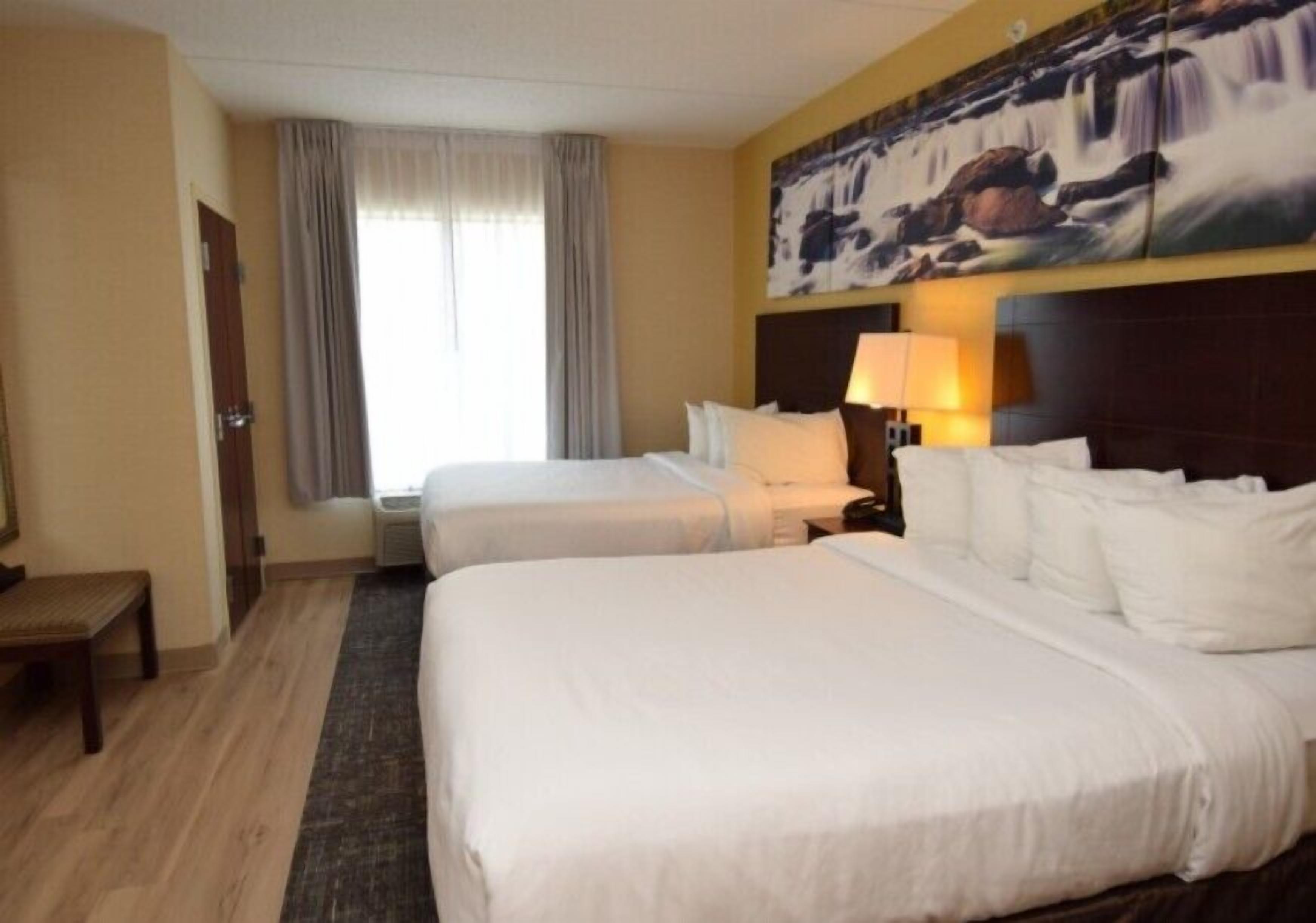 Une chambre d’hôtel avec deux lits, un bureau, une chaise, une fenêtre avec des rideaux et un tableau au mur.