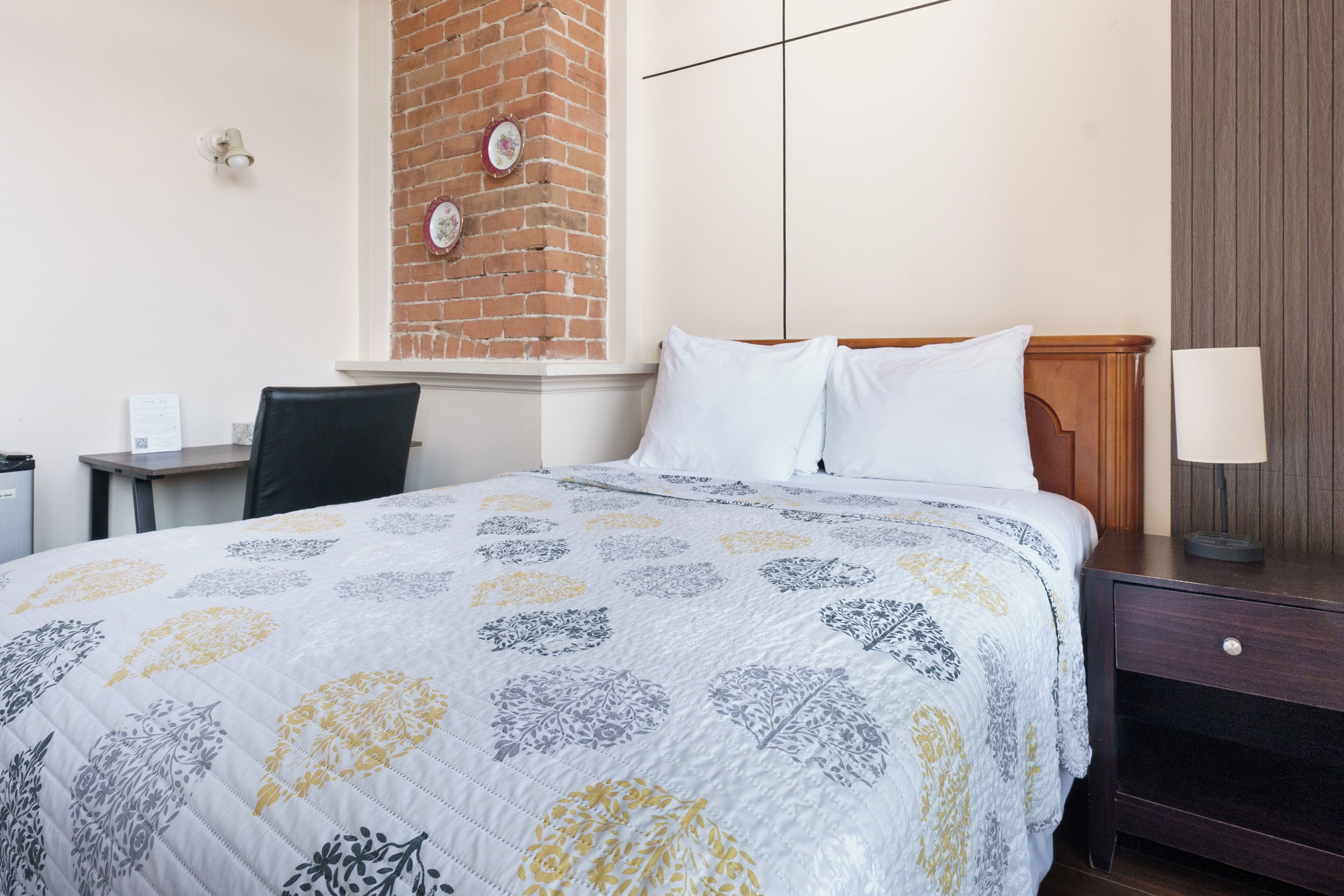 Un lit bien fait, avec une couverture à motifs, une tête de lit en bois, une table de chevet avec une lampe et un mur en briques orné d’assiettes décoratives.