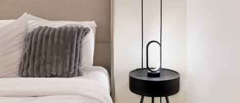 A neatly made bed with white linens and a gray textured throw blanket, a black side table with a modern lamp, and a plain white wall.
