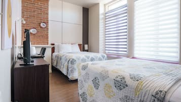 Une chambre d’hôtel avec deux lits, un bureau avec un ordinateur, un mur en briques et de grandes fenêtres équipées de stores.