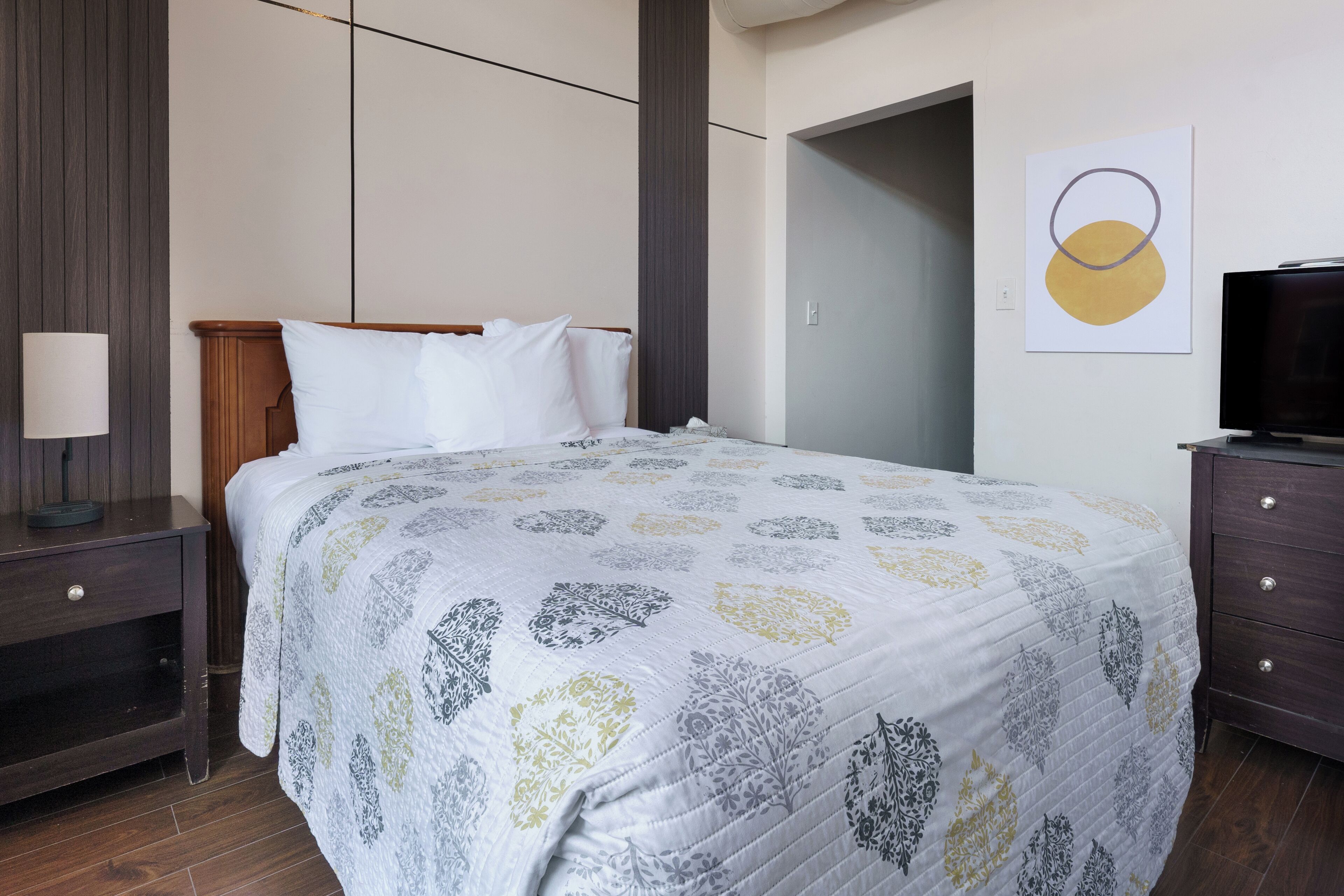 Una cama bien hecha con una colcha estampada, una mesita de noche con una lámpara y un televisor sobre un aparador.