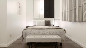 Quarto moderno com cama grande, mesas de cabeceira, um banquinho e luminária fixada na parede.