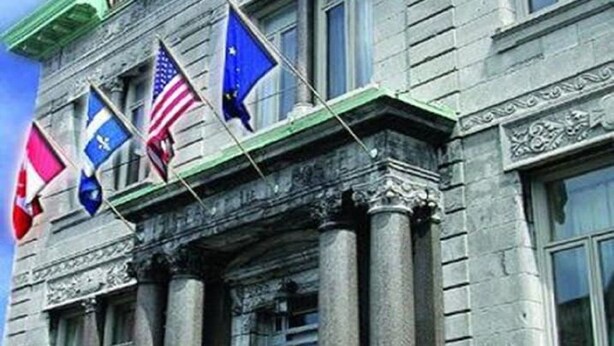 Edifício com estilo arquitetônico clássico, exibindo uma fileira de bandeiras, incluindo a bandeira americana e a da União Europeia, e um telhado verde.
