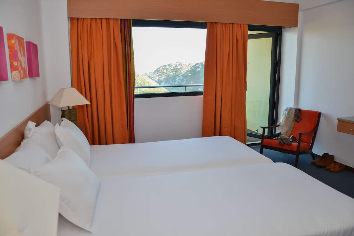 A hotel room with a large bed, orange curtains, a chair, and a view of mountains.