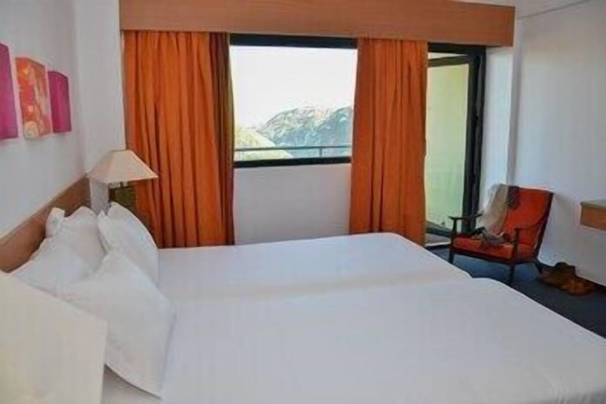 A hotel room with a bed, a chair, a window with curtains, and a view of mountains.