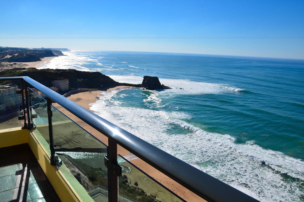 A coastal view from a balcony with a railing, overlooking a beach and ocean.